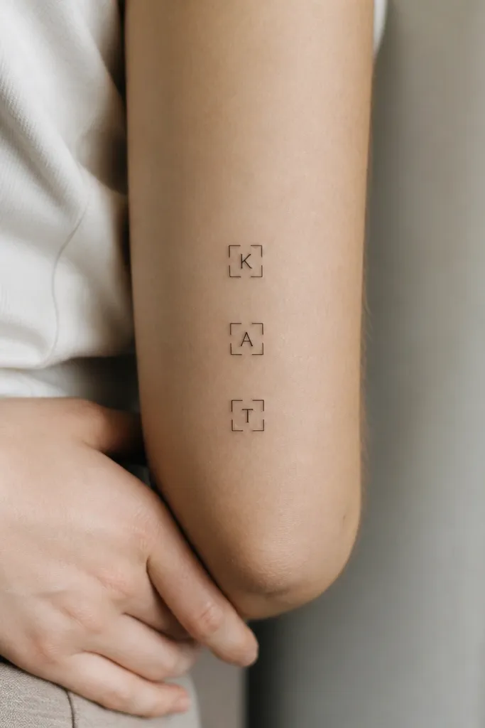 Three small black tattoos of single letters in a monoline font. Each letter sits inside a tiny square bracket frame; the bracket style matches across all three.