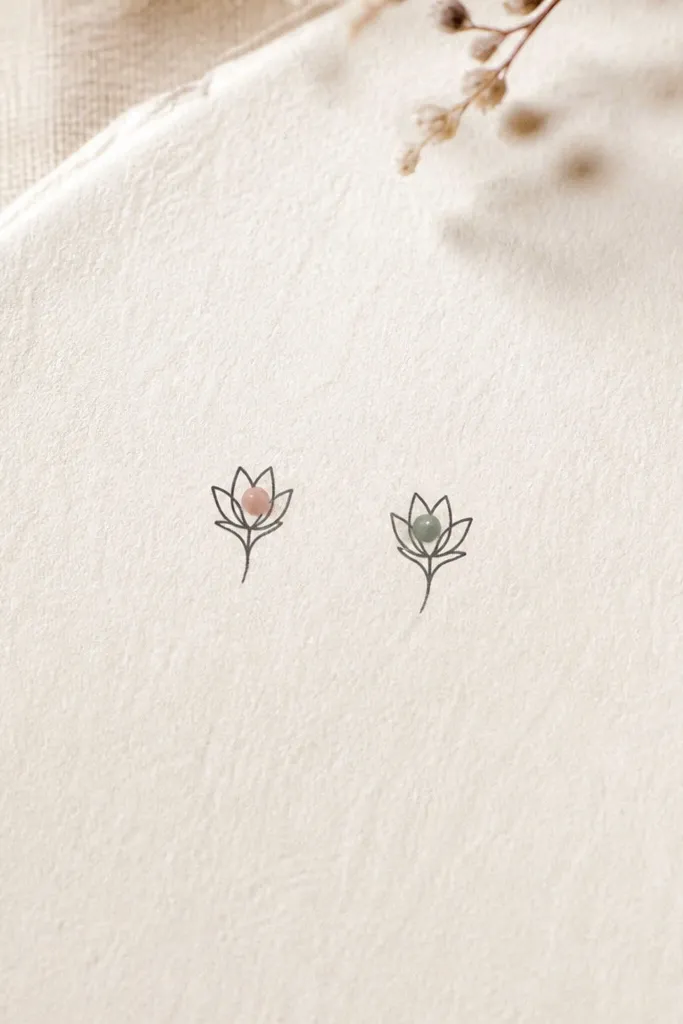 Two matching micro flower bud icons in fine black linework. Each bud has a single colored dot in the center - one tattoo uses soft blush pink, the other uses muted sage green.