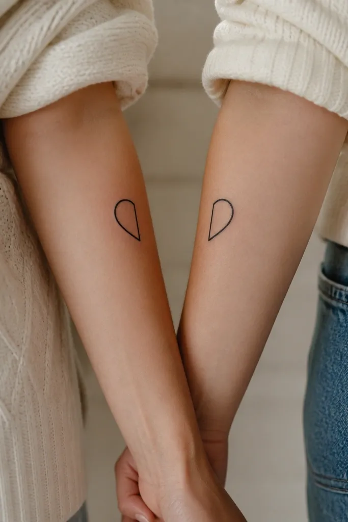 A matching pair of small minimalist half hearts. One tattoo shows a clean black half heart with a flat cut edge; the other shows the matching half heart mirrored so the flat edges meet when both wrists are held together. Both are about palm-sized in negative space and placed on the outer forearms.
