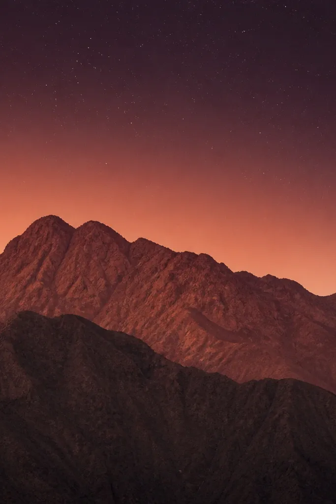 Two overlapping mountain layers in terracotta and cocoa. The sky fades from peach near the horizon into deep plum at the top. A light dusting of micro-stars fills the upper sky.