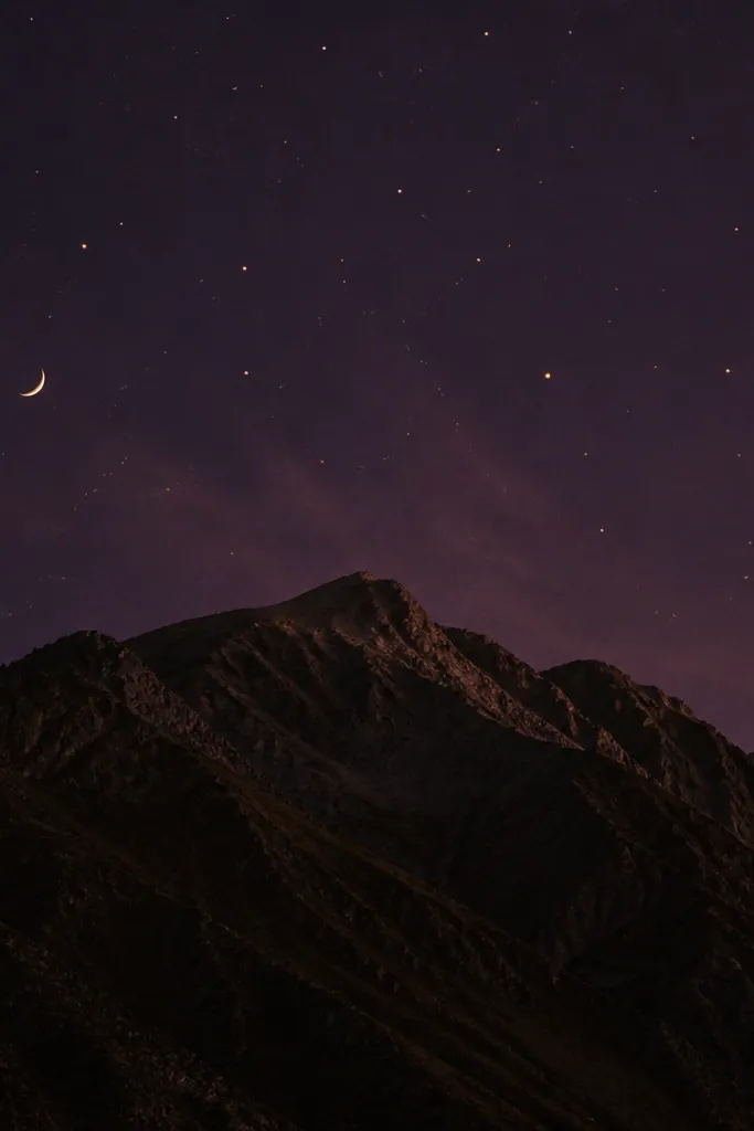 A mostly dark mountain ridge in deep brown and black. Above it, the sky is purple-black with scattered warm ember dots in gold and soft orange. A tiny crescent moon sits off to one side.
