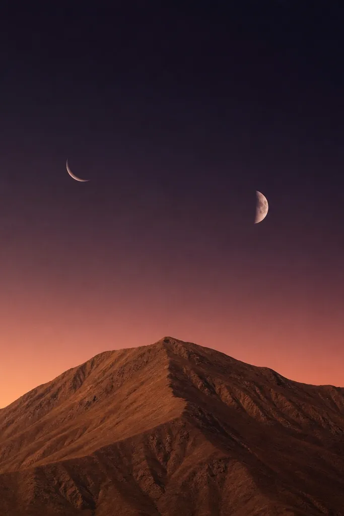 A single warm brown ridge with two small moons above it: one crescent and one partial moon. The sky is deep purple-black with a warm peach gradient near the horizon.