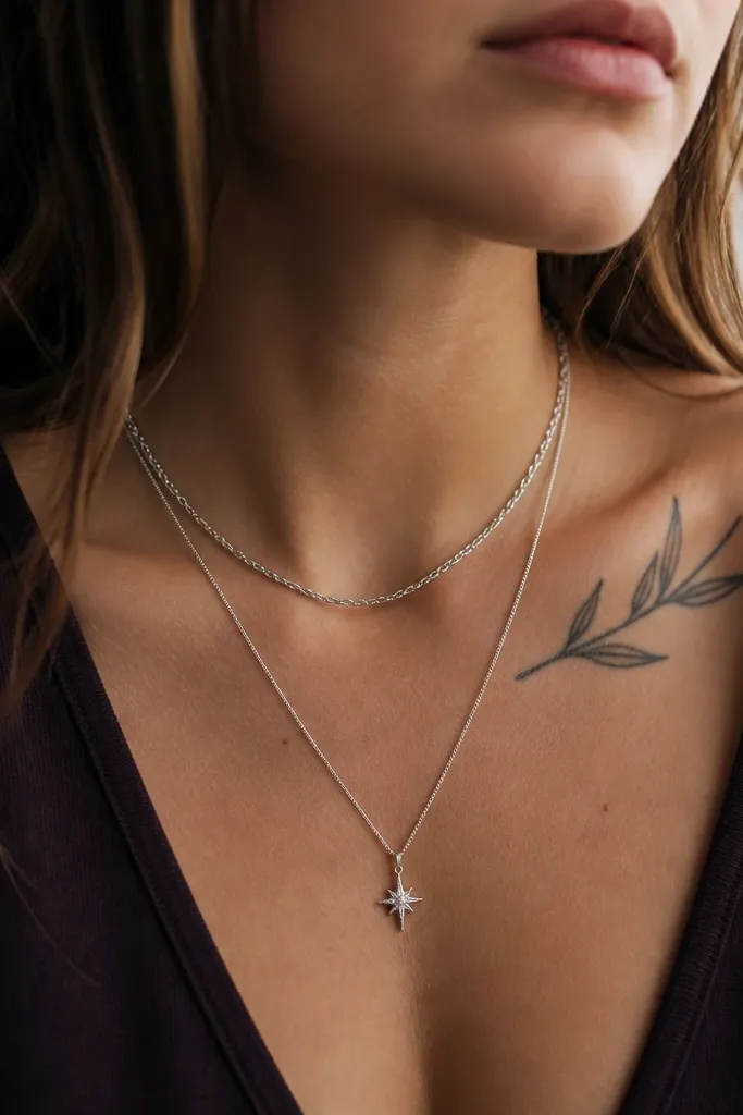 Two necklaces layered: a short silver chain and a longer charm necklace with a small star pendant. The tattoo is visible on the upper chest/neck area, with a dark plum top beneath.