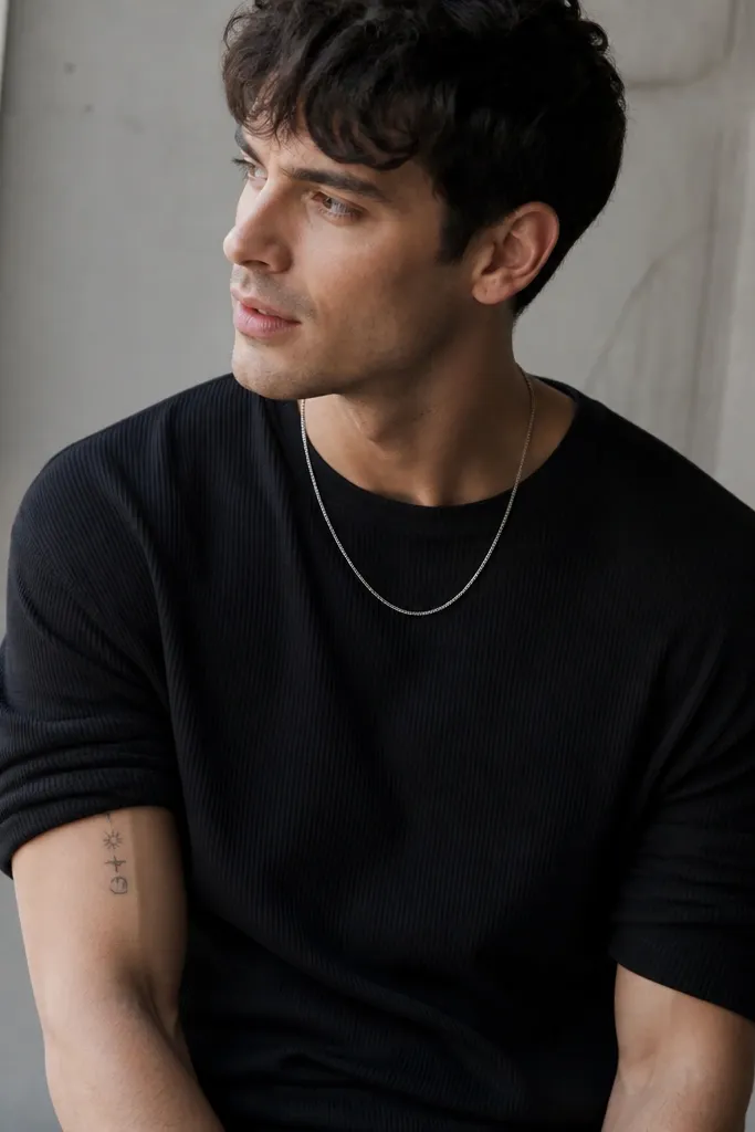 A ribbed black t-shirt worn with the sleeves pushed up, revealing an inner arm tattoo of a small symbol cluster. A plain silver chain necklace sits at the neckline.