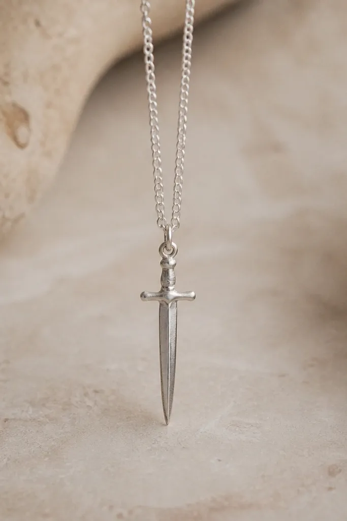 A tiny vertical charm shaped like a slim blade or dagger, with a small crossguard and two thin hanging lines. Shading sits along one side of the blade.