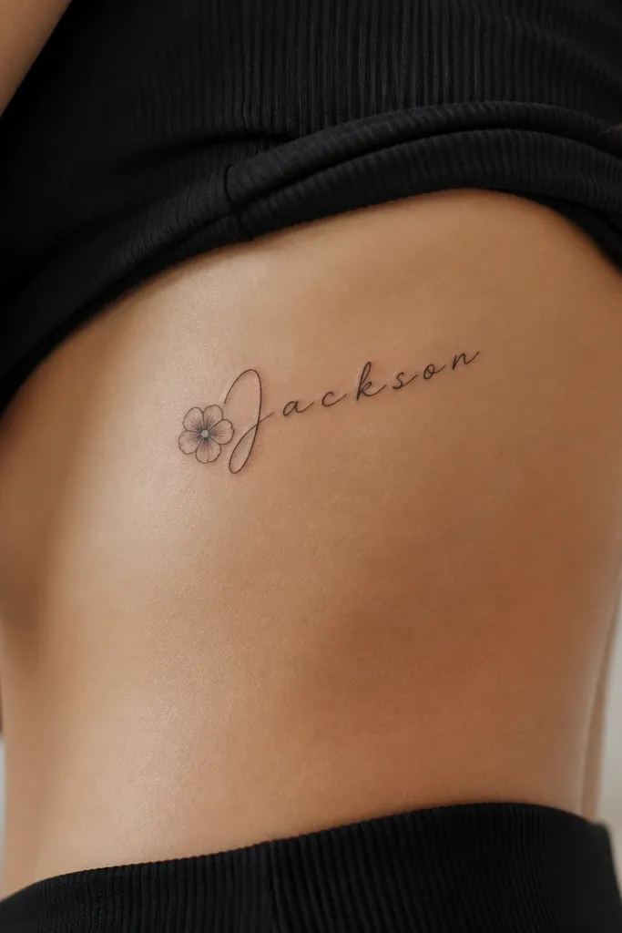 A side rib tattoo: the boyfriend's name in elegant black script, slightly curved, with a tiny five-petal flower next to the first letter. The flower has light stippling shading.