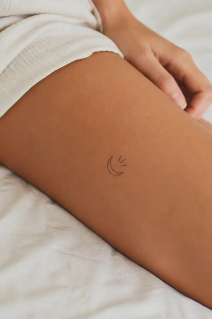 A small crescent moon outline with three tiny straight lines like emphasis marks, placed on the outer thigh above the knee.