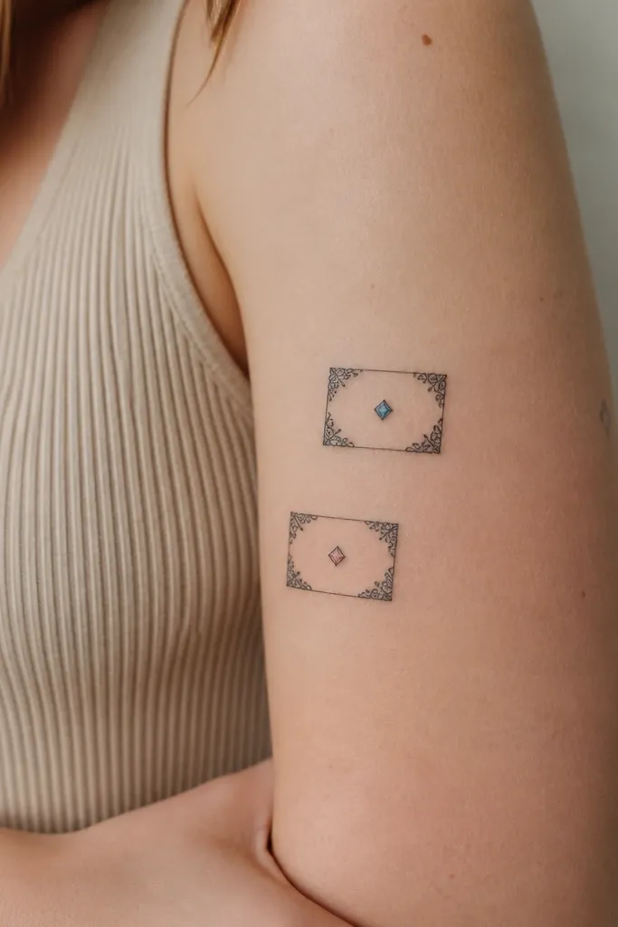 Two small rectangular tattoos with delicate lace-like corner flourishes in black linework. Each frame holds a single tiny gemstone-shaped dot in a muted color (one more blue, one more pink).
