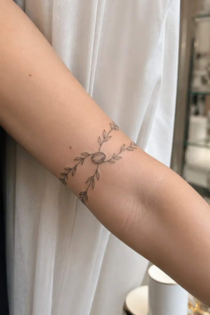 A fine line olive branch tattoo has two slender stems running parallel along the forearm, curving slightly toward each other. They meet at a central olive oval near the middle. Leaves sprout from both stems, with the densest cluster around the central oval.