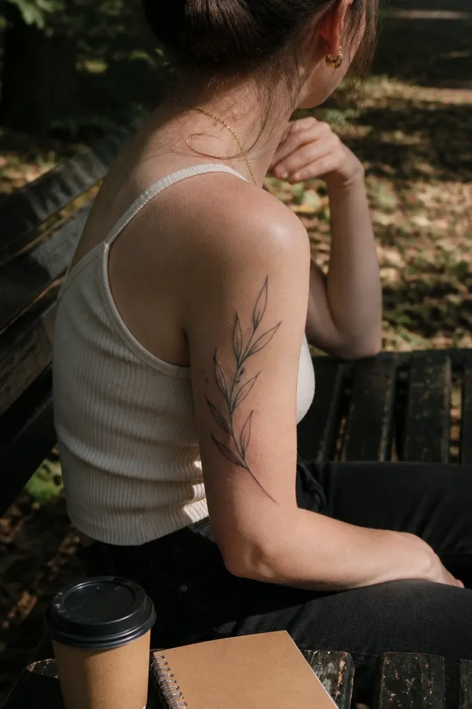 A small olive branch tattoo on the back of the upper arm shows leaves with slightly softened tips, black ink, thin linework, photographed with arm turned.