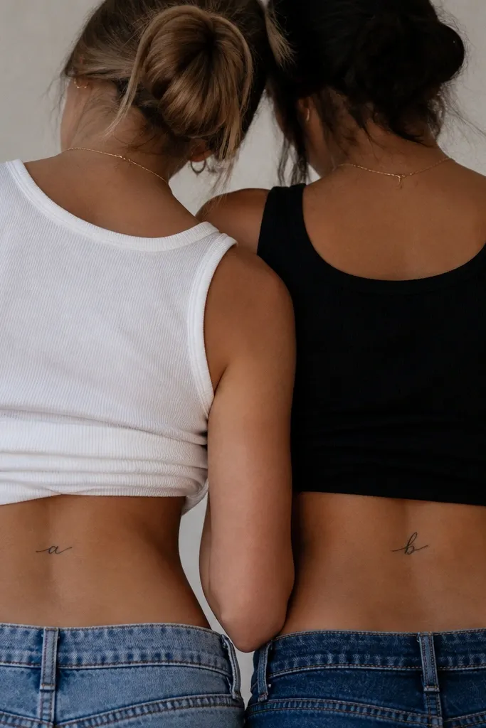 Two matching lower-back tattoos, each a small lowercase script initial in black. The script is thin, flowing, and the letter sits horizontally with clean negative space around it.
